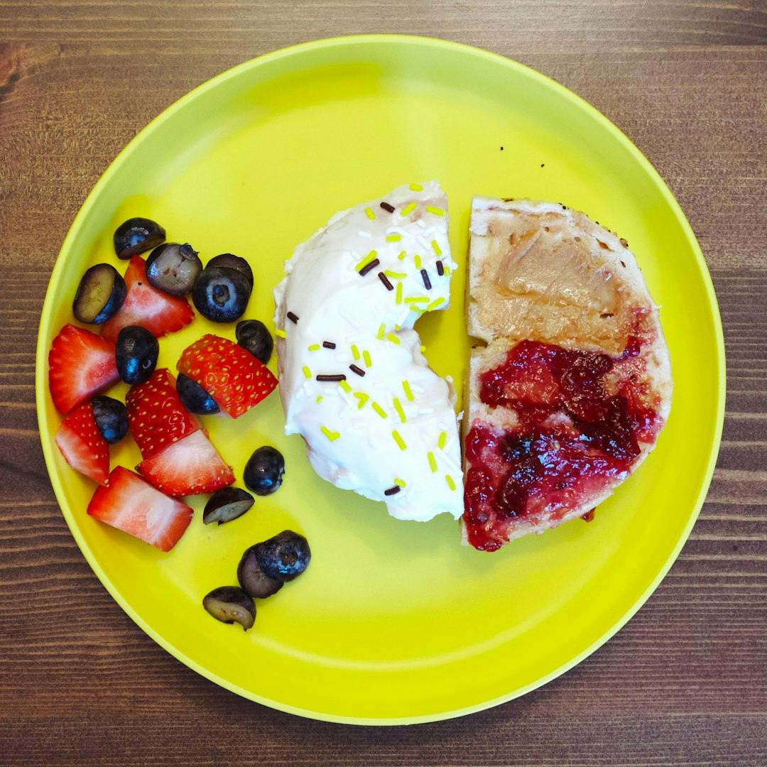 A bright yellow plastic plate with freshly sliced strawberries and blueberries served with a sprinkled donut and bagel with peanut butter and jelly